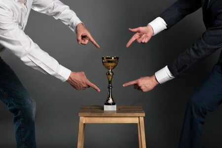 Two businessmans is showing by index fingers on a golden cup award trophy on a chair. Struggle for a...