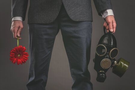 Man is holding in hands a red gerbera flower and gas mask on a gray background. Pollution of...