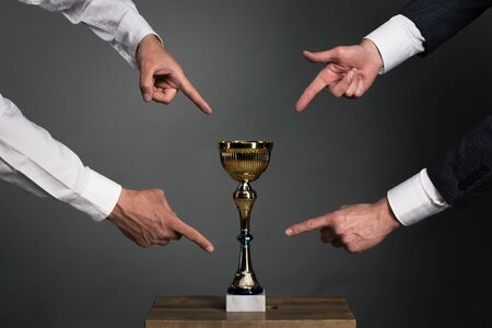 Two businessmans is showing by index fingers on a golden cup award trophy on a chair. Struggle for a...