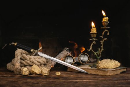 Pirate concept. Treasure hunt. Adventurer table with treasure gold, sword, binoculars and rope on a burning fire background.の写真素材