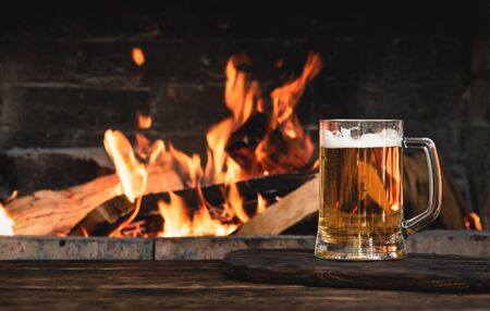 Beer in a mug on a wooden table on a burning fire in a fireplace background.
