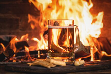 Beer in a mug and cheese sticks on a wooden table on a burning fire in a fireplace background.の写真素材