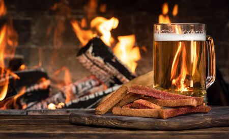 Beer in a mug and a hard chucks on a wooden table on a burning fire in a fireplace background.