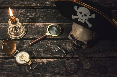 Pirate captain table with pirate hat on the human skull, compass, magnifying glass, mooring rope, coins, key of treasure chest and burning table. In the searching of gold.の写真素材