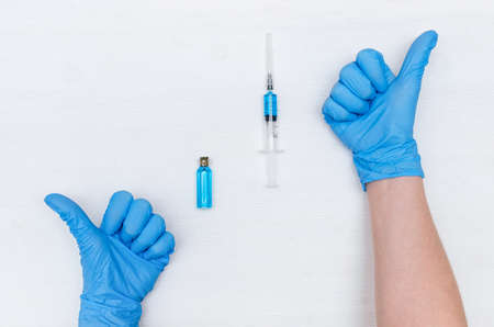 Doctor hands in medicinal gloves showing a thumbs up sign and a syringe with blue liquid medicine on the doctor table background. Choose the best drug concept.の写真素材