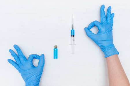 Doctor hands in medicinal gloves showing a ok sign and a syringe with blue liquid medicine on the...