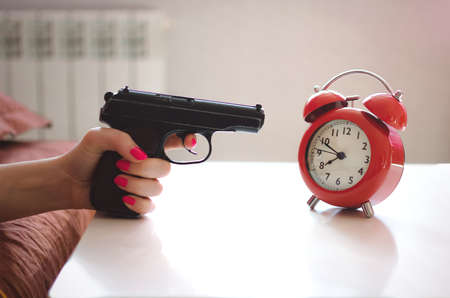 Sleepy woman in the bed is trying to kill a red alarm clock on the table by the gun in her hand. Kill the alarm clock concept.の写真素材