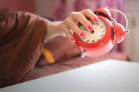 Sleepy woman in the bed is trying to turn off a red alarm clock on the table at morning time.の写真素材