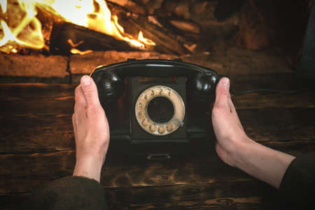 Man is holding in hands a black old phone on a burning fire in fireplace background. Hotline concept.の写真素材