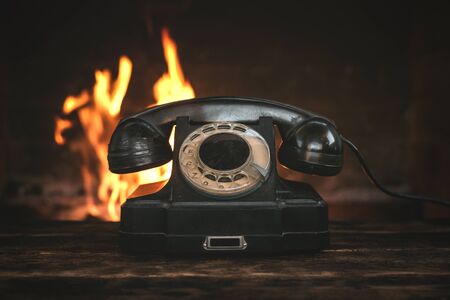 Black old phone on a table on a burning fire in fireplace background. Hotline concept.の写真素材