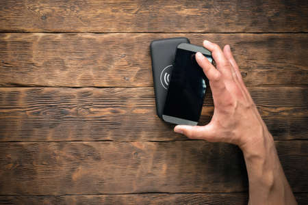 Wireless charger and mobile phone in male hand on a wooden table background.の写真素材