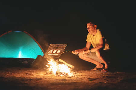 Tourist is cooking a sausages on the campfire on tent background at night.