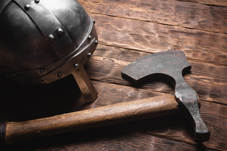 Old viking ax and iron helmet on the brown wooden table background close up.の写真素材