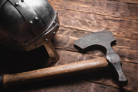 Old viking ax and iron helmet on the brown wooden table background close up.