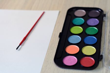 Palette of watercolors, piece of white paper and a brush on a wooden tableの写真素材
