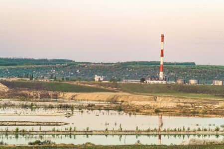 Natural lake is a chalky quarry with a factory smokestack under a green hill on the horizon....