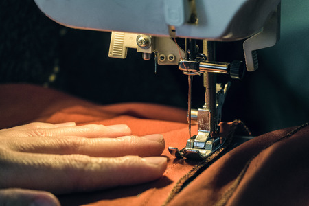Seamstress sews clothes made of red cloth on a sewing machine. Work by the light of the built-in...