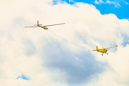 Yellow small light-engine sport plane flying in the cloudy sky pulls on a rope the glider planeの写真素材