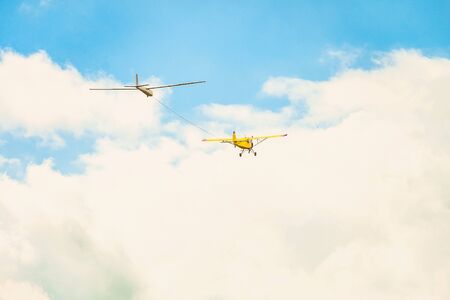 Flying in a bright cloudy sky, a yellow small sports plane with a light engine, pulls the rope by the glider plane.の写真素材