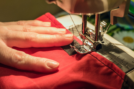 Seamstress sews clothes made of red cloth on a sewing machine. Work by the light of the built-in...