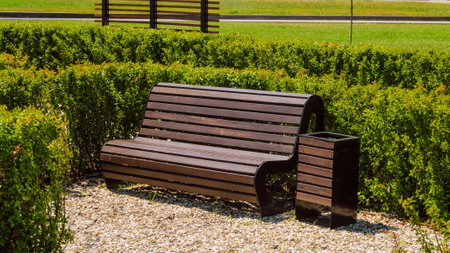 Brown wooden bench and trash can in square with green shrubs. City street improvement and...