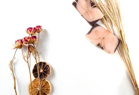 Flat lay composition with two handmade soaps of brown-pink color with an orange tint, dried slices of oranges, roses flowers and spikes of wheat on a white background with wooden texture.の写真素材