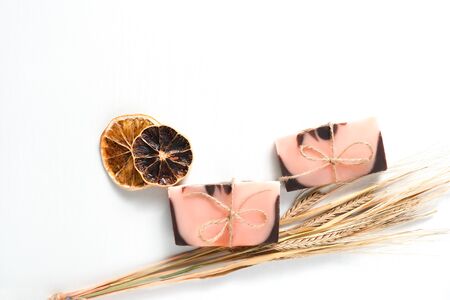 Flat lay composition with two handmade soaps of brown-pink color with an orange tint, dried slices of oranges with a spikes of wheat on a white background with wooden texture. Objects located in the right part of frame. Horizontal photo, copyspace for text. . Handmade cosmetics concept with natural ingredients for personal care, bath, shower and spa.の写真素材