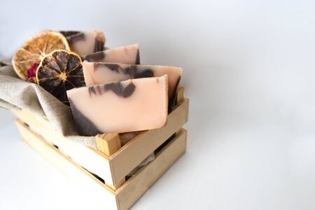 Five bars of handmade soap of brown-pink color with an orange tint , dried slices of oranges and a dried rose in a wooden box on a white background with wooden texture. Horizontal photo, copyspace for text. Handmade cosmetics concept with natural ingredients for personal care, bath, shower and spa. Gift set handmade cosmetics. Selective focus.の写真素材