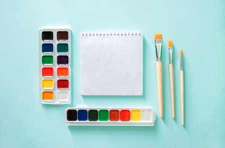 Two sets of color Watercolor paints, three light beige brushes with orange pile and wooden texture and white clear sheet of note book on a light blue background.の写真素材
