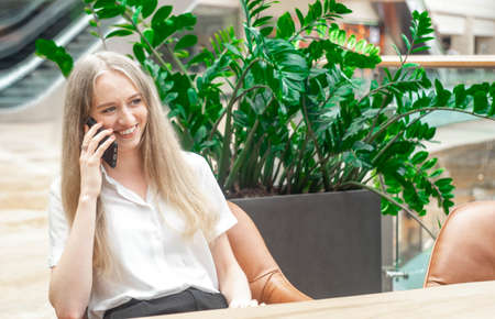 Portrait of a cheerful young caucasian blond business woman sitting on the table, talking at phone and smiling. Concept of successful young women in a corporation. Concept of successful young women in a corporation. Girl in a white shirt. Horizontal photo. Blurred background. Plants on the background.の写真素材