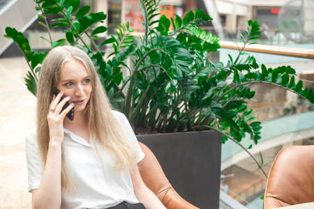 Portrait of a cheerful young caucasian blond business woman sitting on the table, talking at phone and smiling. Surprised face. Concept of successful young women in a corporation. . Girl in a white shirt. Horizontal photo. Blurred background. Plants on the background.の写真素材