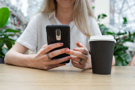Cropped photo of cheerful young caucasian blond business woman in a white shirt typing on smartphone while resting on coffe break. Lifestyle concept. Concept of successful young women on coffe break. Nearby is a black cup of coffee.の写真素材
