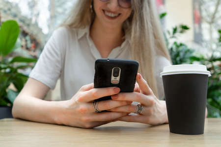 Cropped photo of cheerful young caucasian blond business woman in a white shirt typing on smartphone while resting on coffe break. Lifestyle concept. Concept of successful young women on coffe break. Nearby is a black cup of coffee.の写真素材