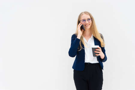 Portrait of a young European blonde business woman is standing in front of a white wall, smiling, talking at the phone and holding coffee in her hand. She is dressed in black pants, blue jacket and a white short-sleeved shirt. Concept of successful young women. Lifestyle concept. Copy space. Space for text. Horizontal photo.の写真素材