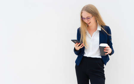 Portrait of a young European blonde business woman is standing in front of a white wall, smiling, looking at the phone and holding coffee in her hand. She is dressed in black pants, blue jacket and a white short-sleeved shirt. Concept of successful young women. Lifestyle concept. Copy space. Space for text. Horizontal photo.の写真素材