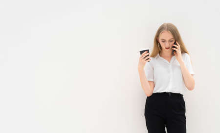 A portrait of young caucasian blonde business woman is standing in front of a white wall, talking to the phone, looking down, thinking and holding coffee in her hand. She is dressed in black pants and a white short-sleeved shirt. Concept of successful young women. Lifestyle concept. Copy space. Space for text.の写真素材