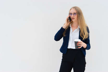 Portrait of a young European blonde business woman is standing in front of a white wall, smiling, talking to the phone and holding coffee in her hand. She is dressed in black pants, blue jacket and a white short-sleeved shirt. Surprised facial expression. Concept of successful young women. Lifestyle concept. Copy space. Space for text. Horizontal photo.の写真素材