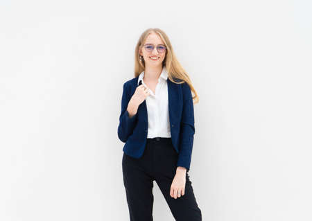 A portrait of young caucasian blonde business woman in glasses standing in front of a white wall, smiling and looking away. The right hand holds a jacket. She is dressed in black pants, blue jacket and a white short-sleeved shirt. Concept of successful young women. Lifestyle concept. Copy space. Space for text.の写真素材