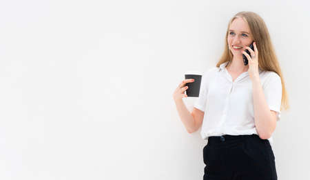 Portrait of a young European blonde business woman is standing in front of a white wall, smiling,...