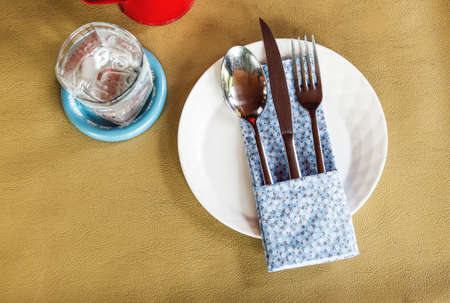 Folded napkin with fork-spoon-knife on white plate on food table with a glass of waterの写真素材