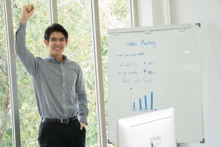 The handsome Asian man, aged 25-35, is standing, raising his fists and smiling happily. He is the...