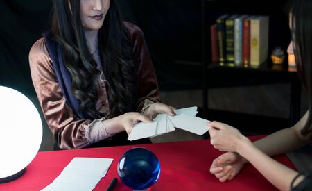 The fortune-telling woman is predicting the fortune for A beautiful Asian woman by a gypsy card. It is a ritual performed in a dark room.