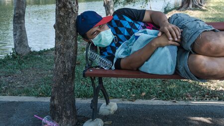 Asian homeless man wearing protective mask sleeping on a bench. Unemployed due to the impact of the spread of the virus.の写真素材