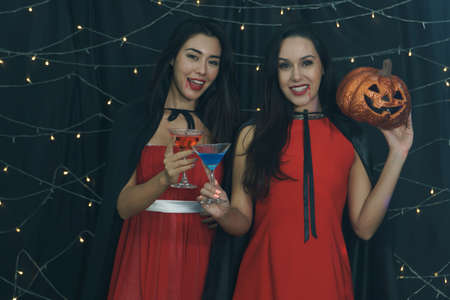 Beautiful woman and friend in Halloween devil costume with carved pumpkin and drinking cocktail. Have fun at a party in nightclub decorated with lights. Halloween celebrationの写真素材