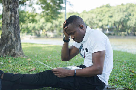 African American young man reading news on his laptop in green forest park.African business young man in casual clothes working on laptop on green grass. muscular young man learning online on a laptopの写真素材