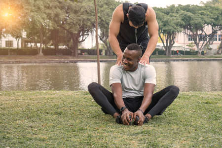young man African-American and Asian male friend talk about hilarious health on the lawn in green forest park. Athlete outdoor exercise. Sportsman warm up and stretches. trainer relax after training.の写真素材