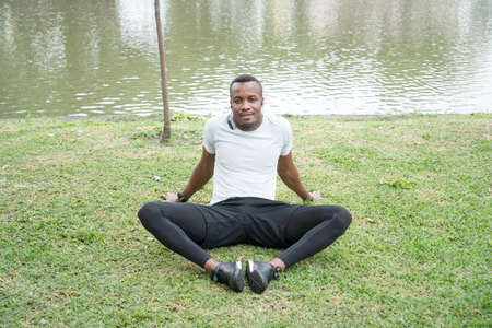 Happy smiling positive African man in sportswear it down and listen to music on the green grass. Athlete male relax after exercise outdoors. Runner stretching and warm up before training.の写真素材