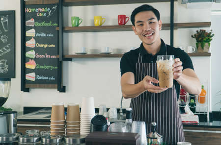 Asian young man barista preparing coffee in cafe. waiter holds an Iced Latte cup and hand it to the customer at front counter. owner of coffee shop happy smiling in business successful.の写真素材