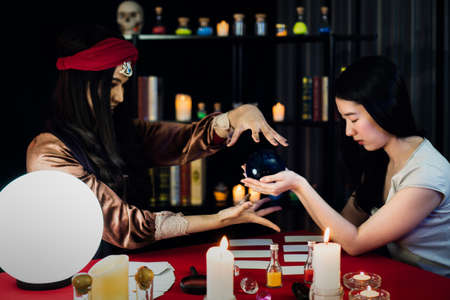 attractive beautiful woman with fortune teller female Ritual luck zodiac Prophecy Mystery crystal ball. lady dressed in witch Halloween magic trick in a dark room candle light bokeh.の写真素材