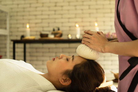Close up Asian young face beautiful woman nature forehead massage with hot compress in spa salon. Traditional therapist is giving herbal Thai massage for female relaxing in beauty spa.の写真素材